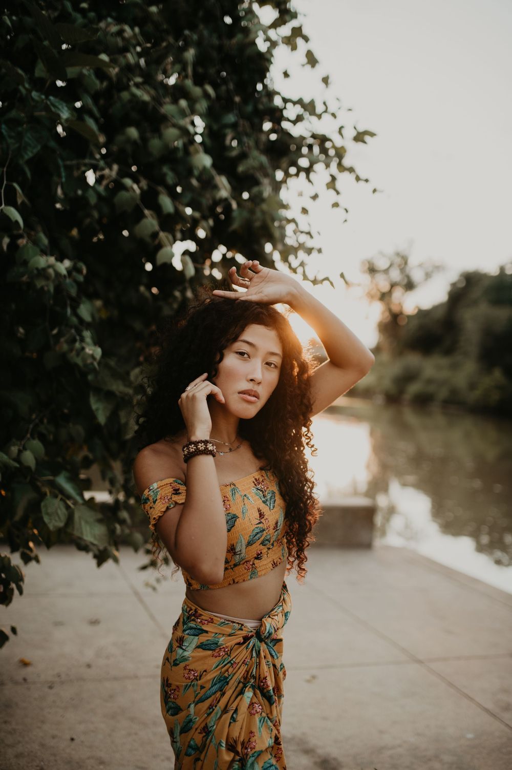 Person in floral dress poses gracefully by river during golden hour.