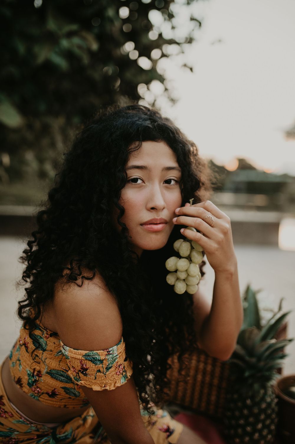 A dreamy portrait showcasing long curly hair and a floral off-shoulder dress while holding grapes.