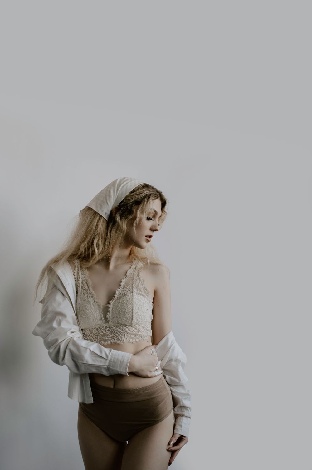 Soft portrait of someone in intimate white lingerie and oversized shirt against grey backdrop.