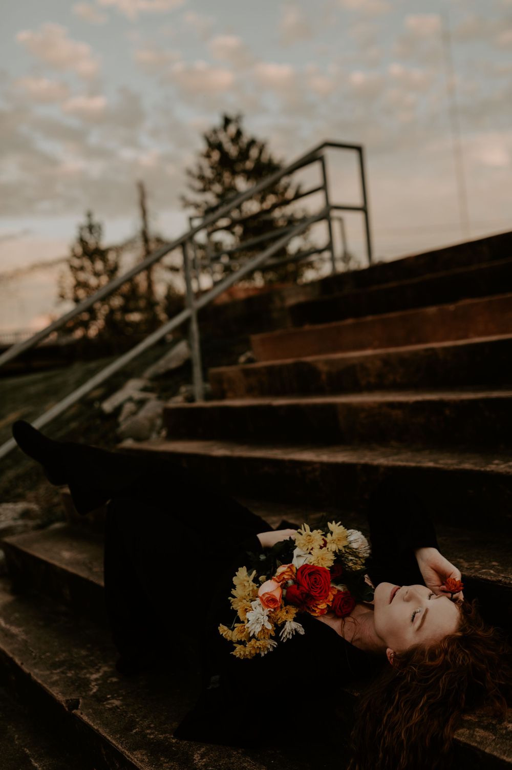 A dreamy portrait of a person lying on wooden steps with colorful flowers against a moody sky.
