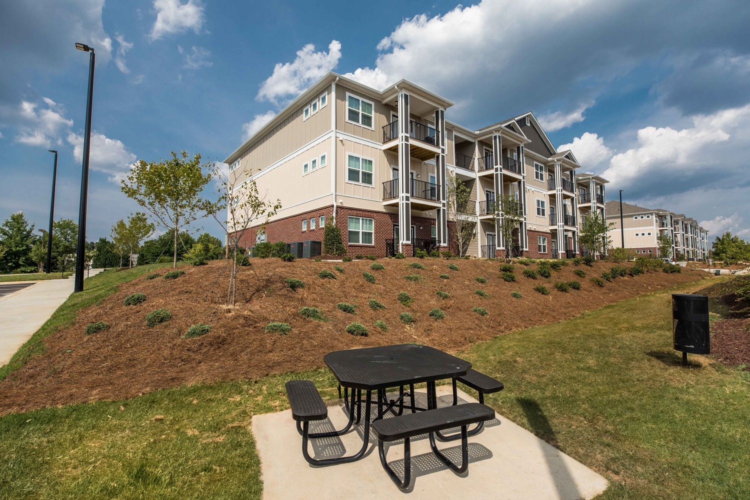 Modern apartment complex with picnic table on landscaped hill under blue sky.