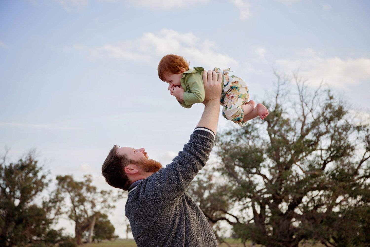 father holding baby daughter in the air outside smiling at each other