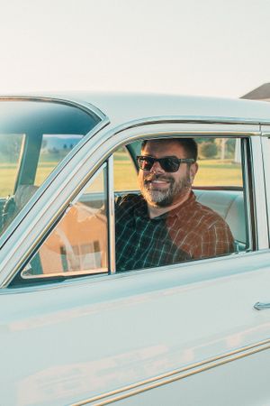 Classic vintage car with driver wearing sunglasses and plaid shirt on sunny day.