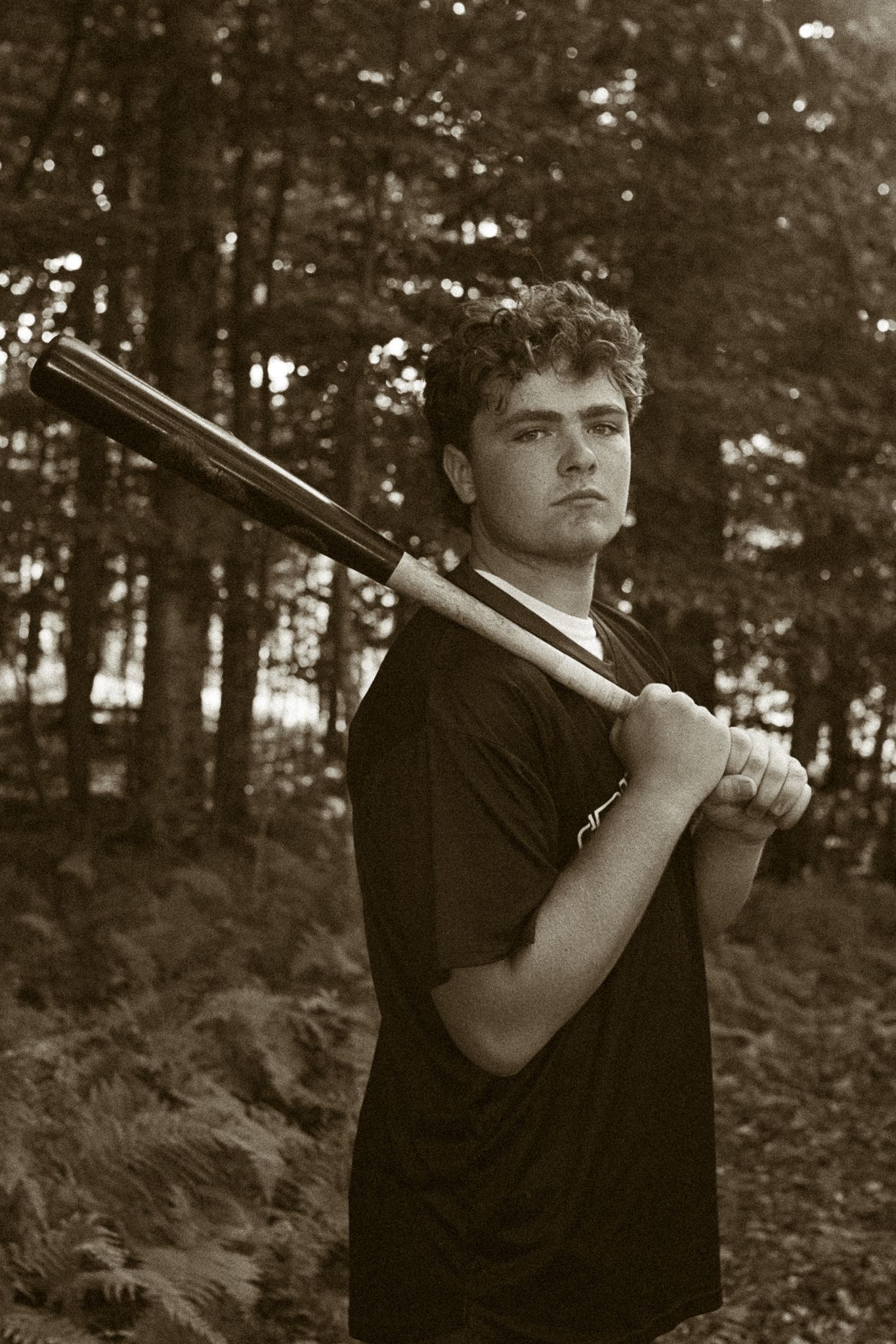 Dramatic black and white portrait of someone holding baseball bat in wooded setting.