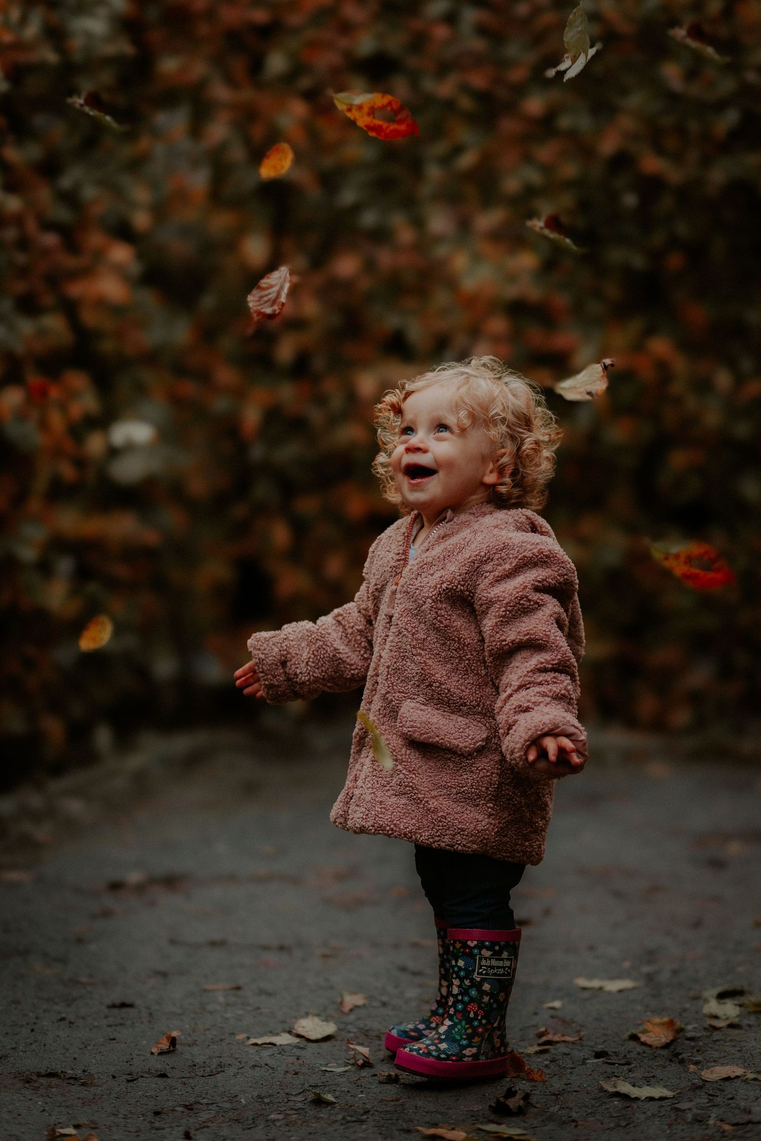 Joyful toddler in a fluffy pink coat and floral boots plays with falling autumn leaves on a moody afternoon.