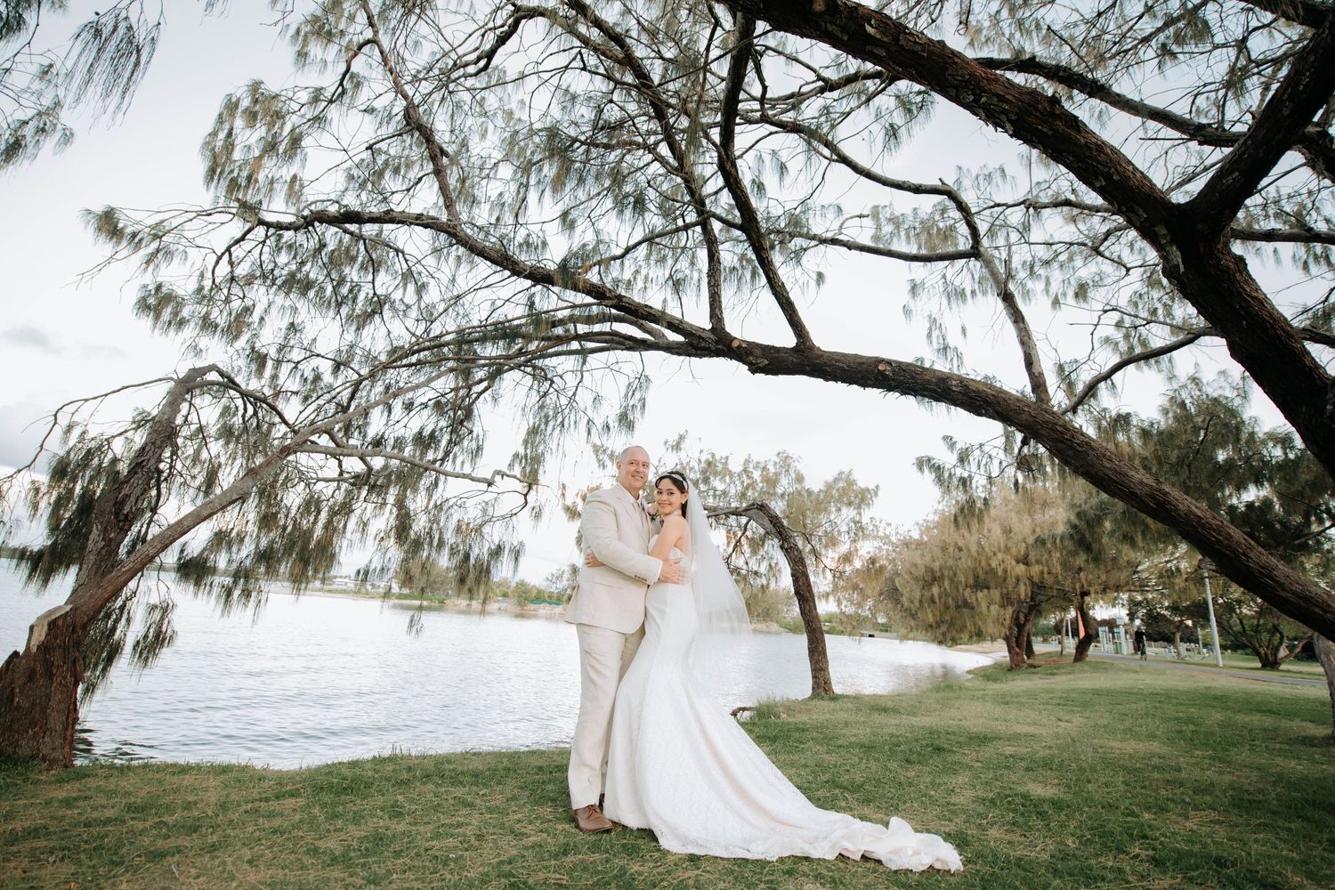 Wedding couple poses under willow trees near waterfront in elegant attire.