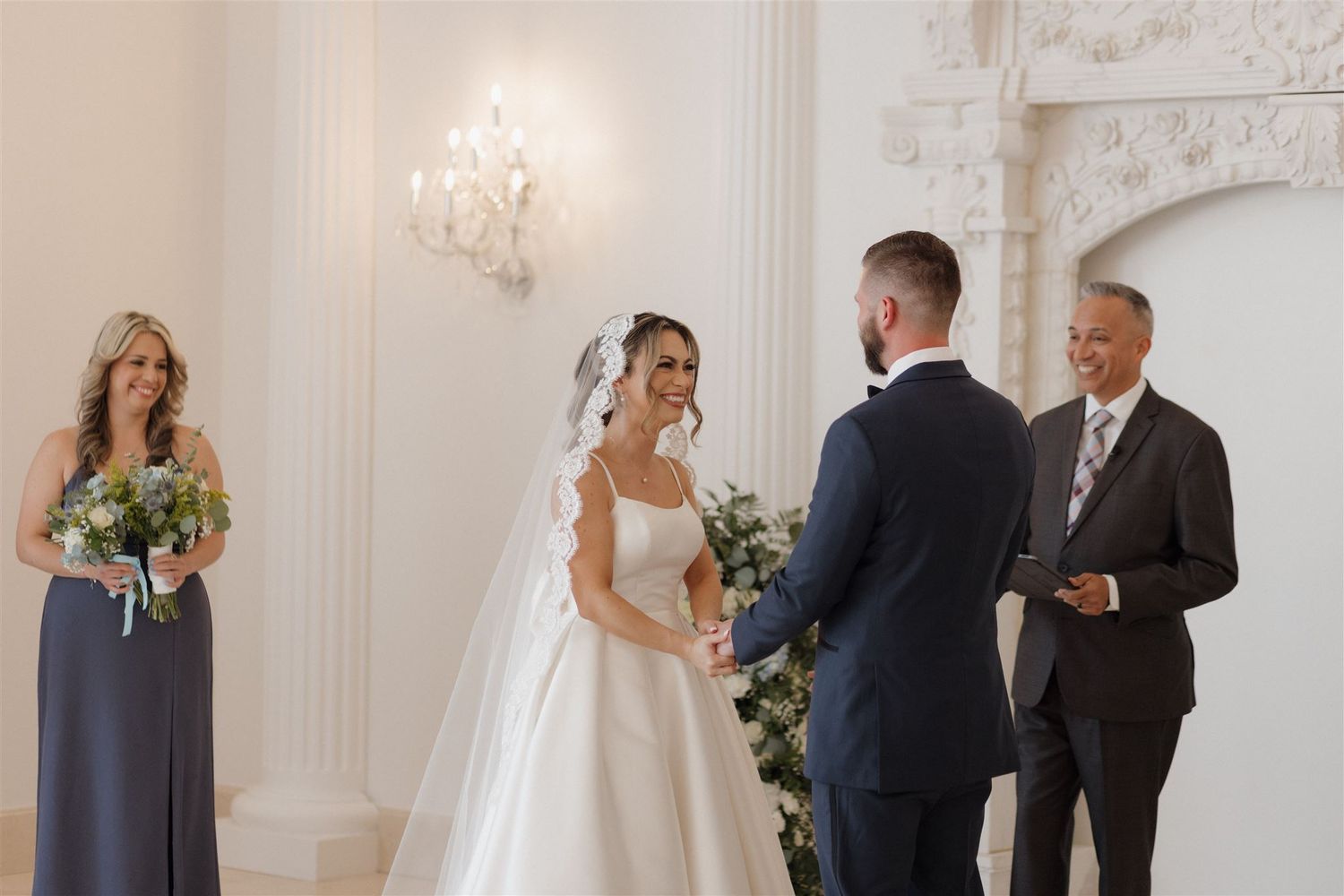 A wedding ceremony takes place in an elegant white room with crystal chandeliers and ornate architectural details.