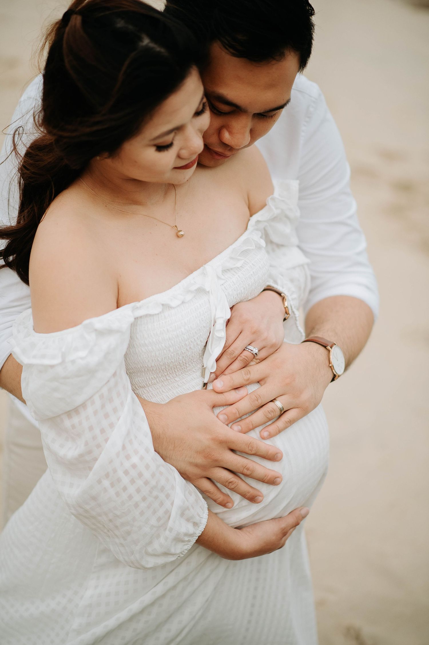 A tender maternity moment captured on the beach in white attire.