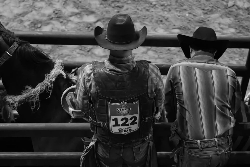 Cowboys in hats lean against a railing watching an event in black and white.