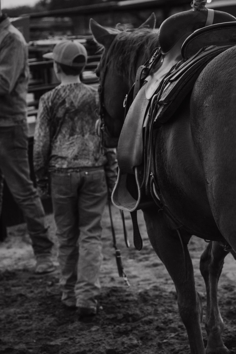 Black and white view of people and horses at a riding event, focusing on saddles and legs.