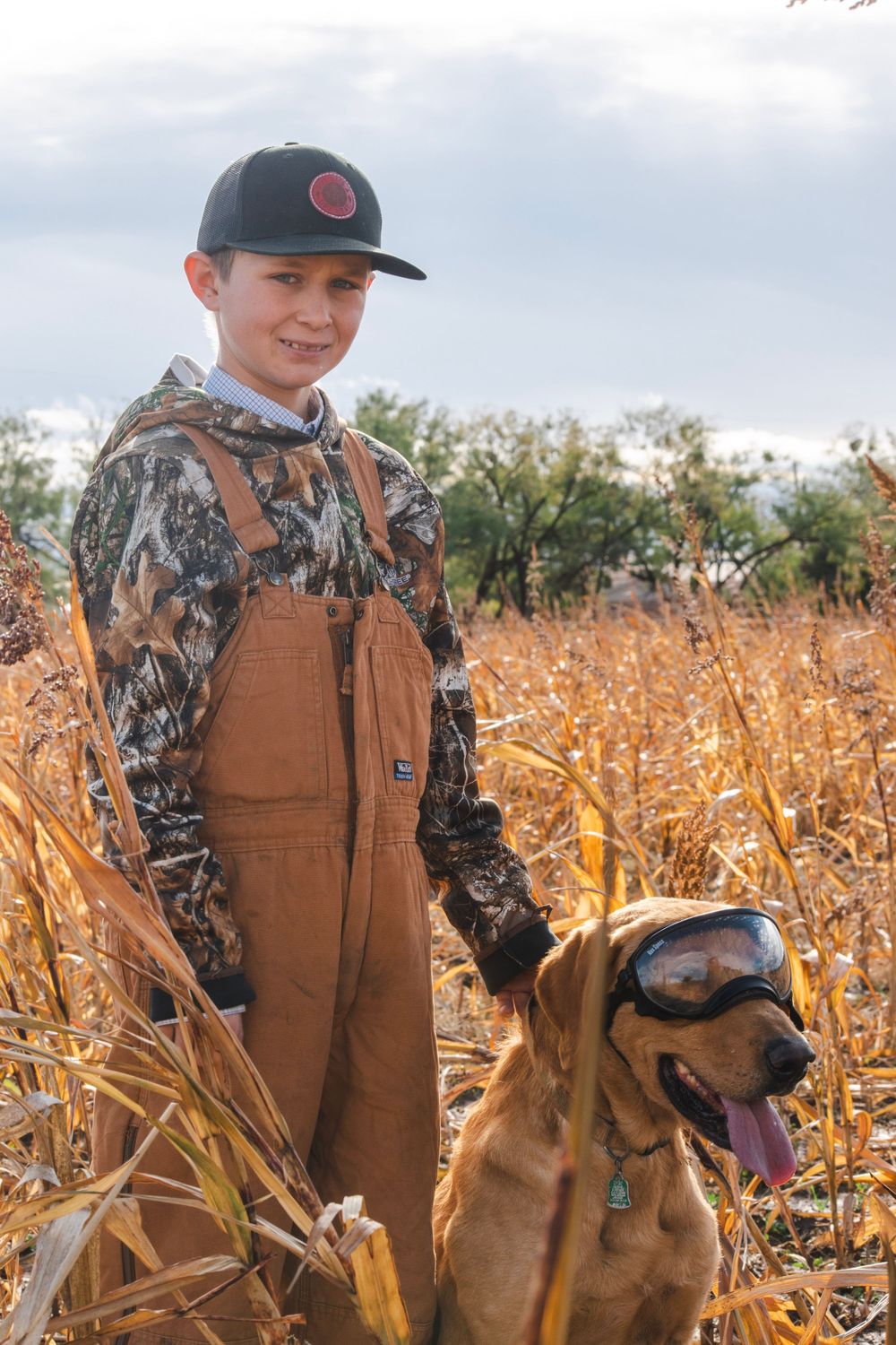 Dog and handler in brown overalls stand in autumn cornfield during hunting season.