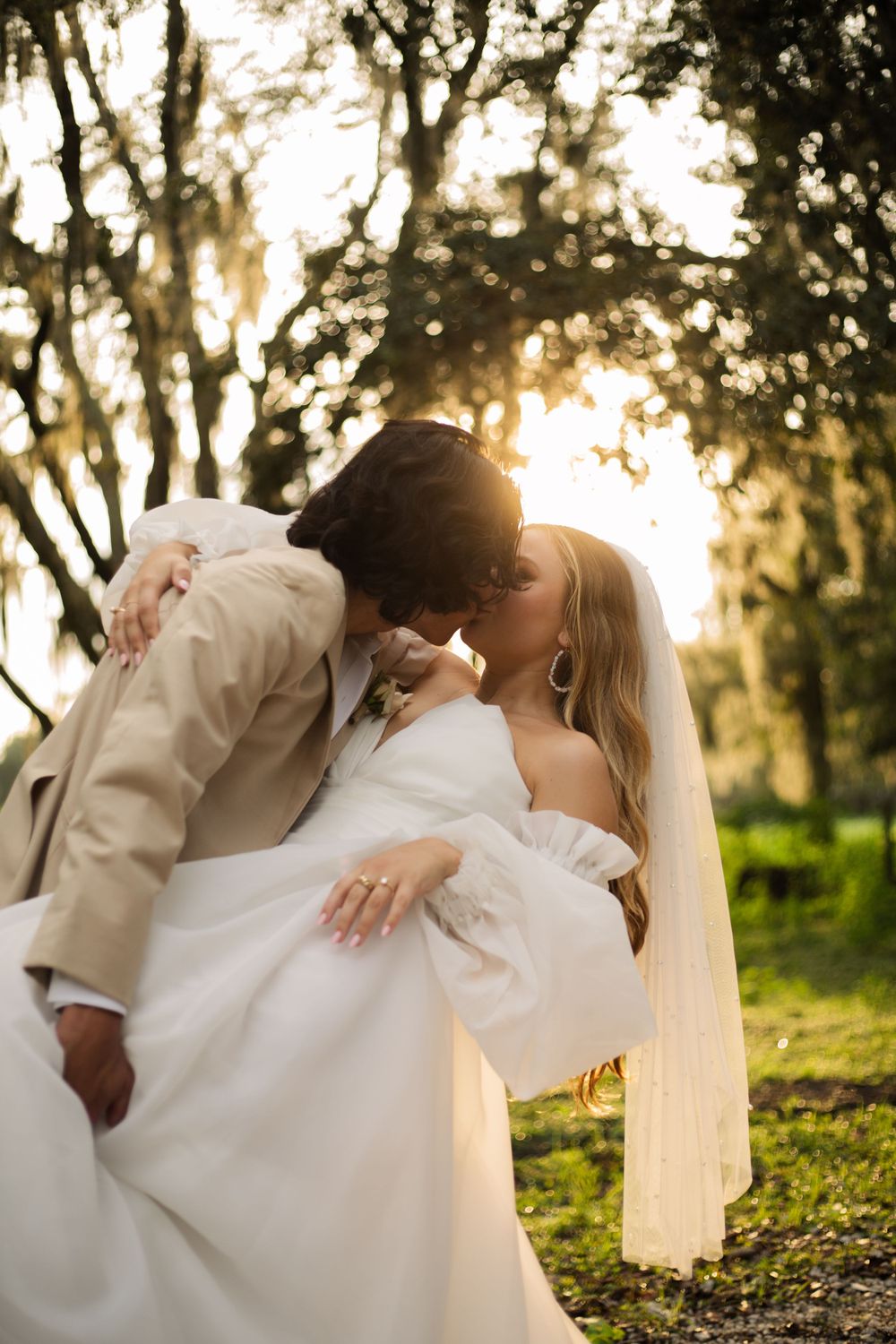 Couple in wedding attire shares a romantic dip pose against a backlit forest background.