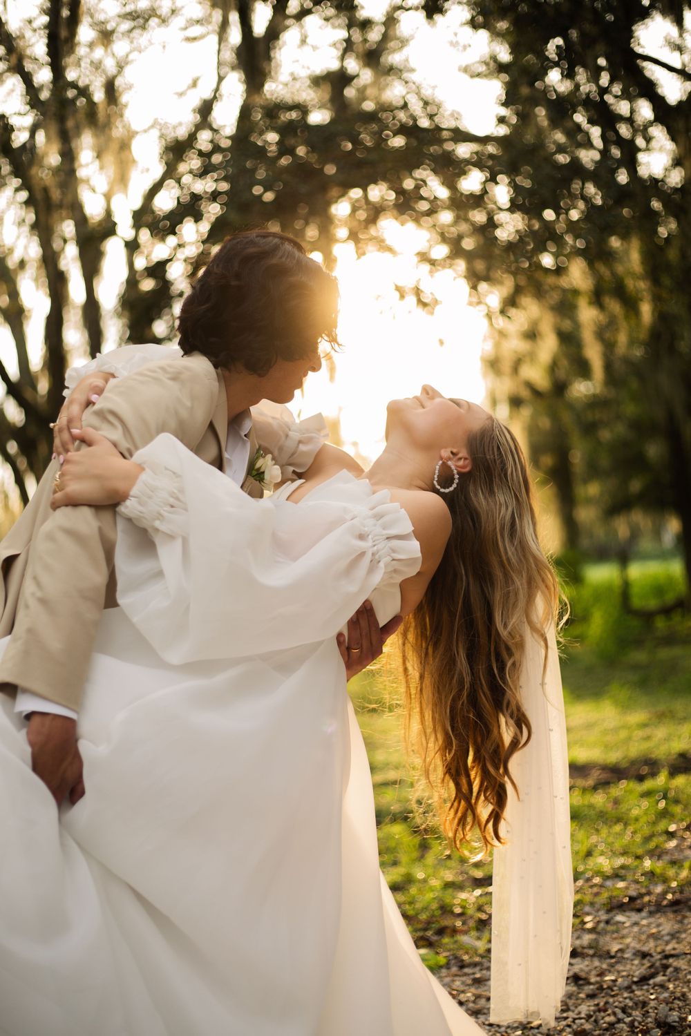 Elegant wedding portrait with dramatic dip pose under sunlit trees with flowing veil.