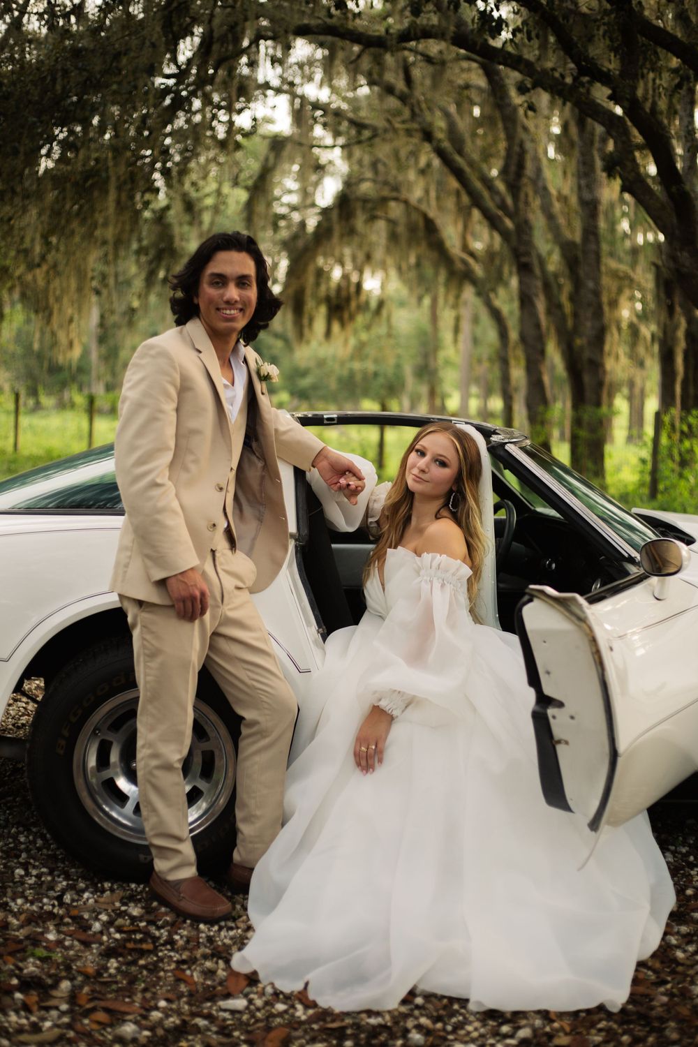 Stylish wedding couple poses with white vintage car under moss-draped trees.