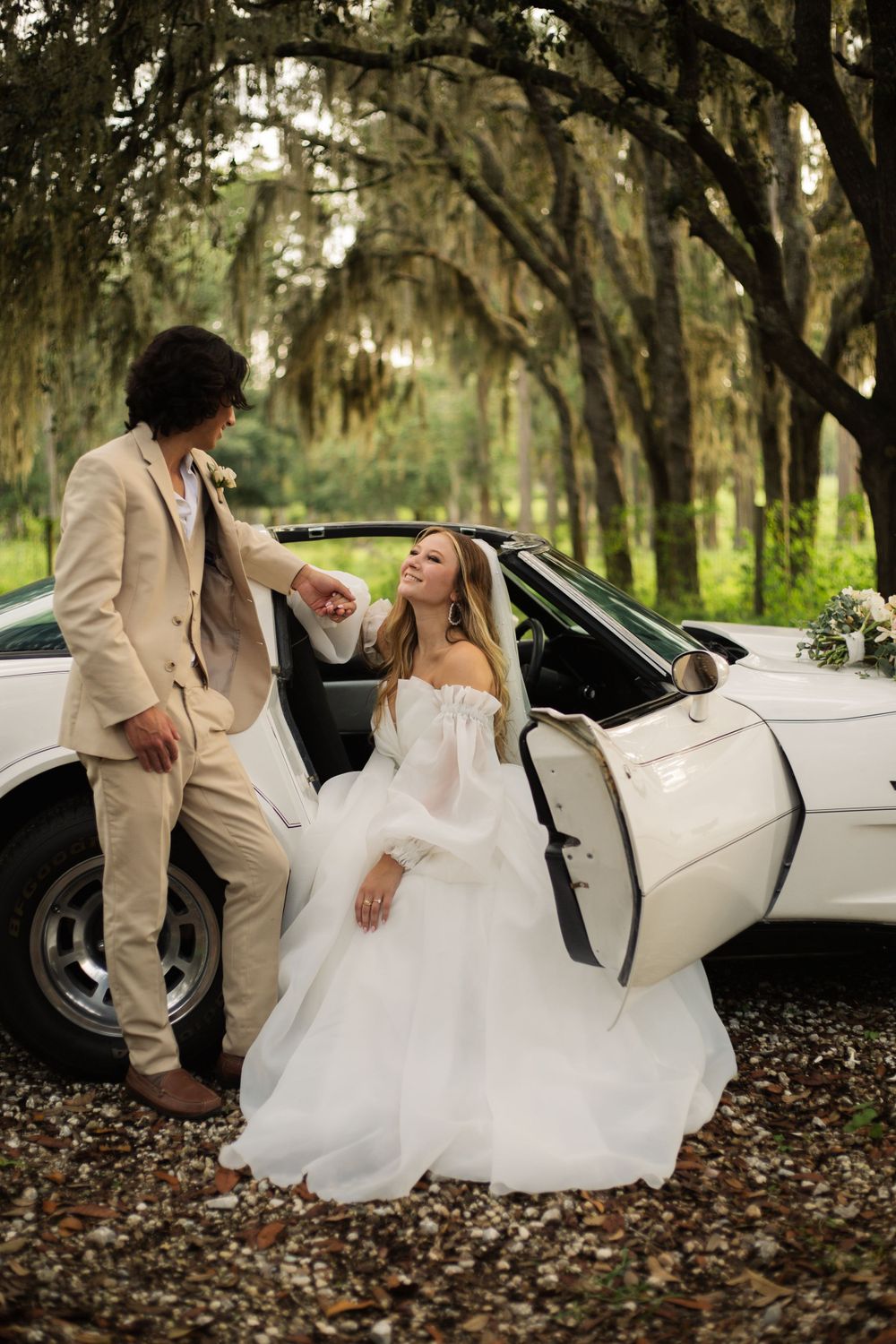 Wedding couple shares a playful moment by their white vintage car on a tree-lined path.