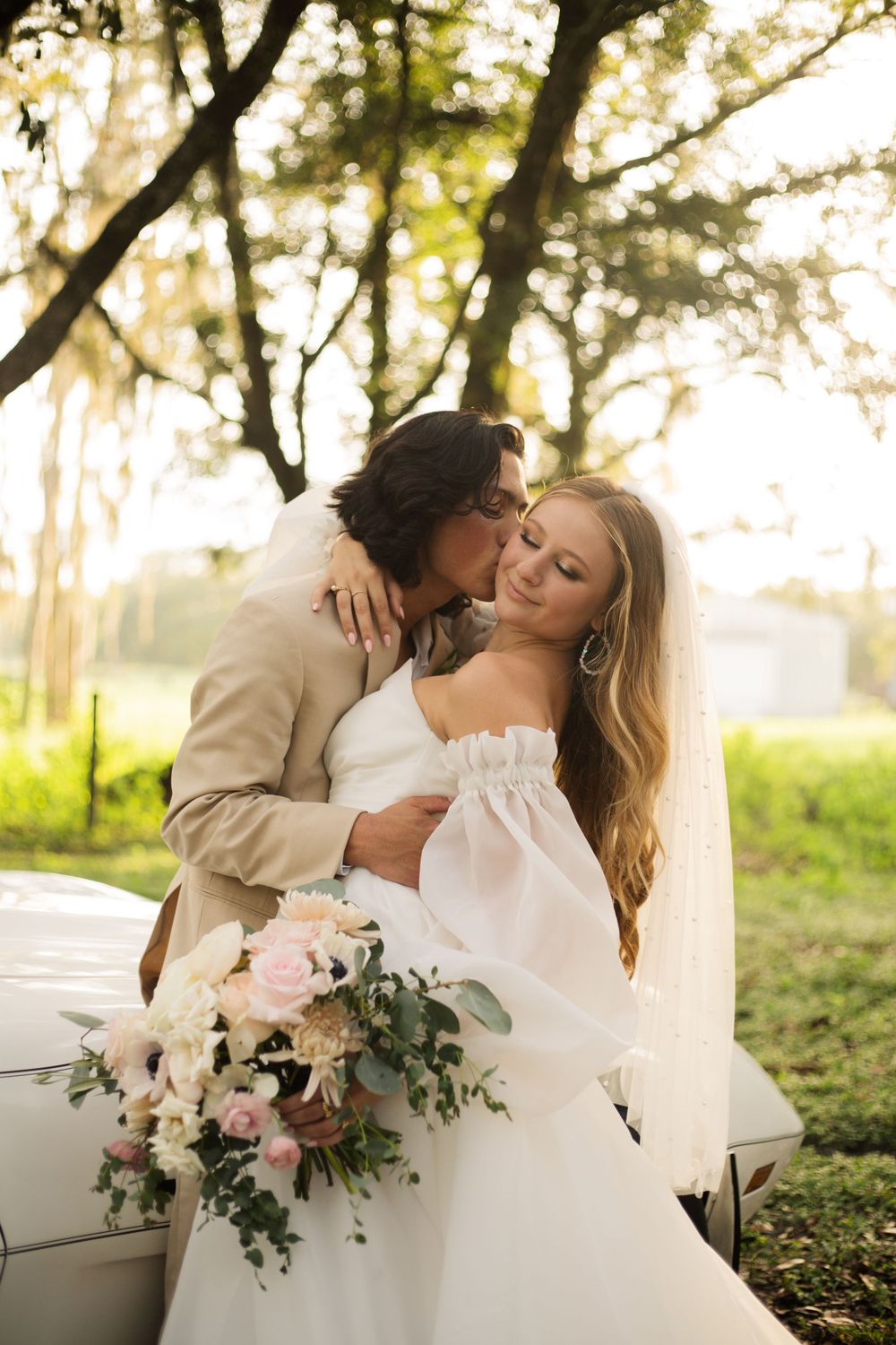 Romantic sunset wedding portrait with a vintage white Corvette and floral bouquet.
