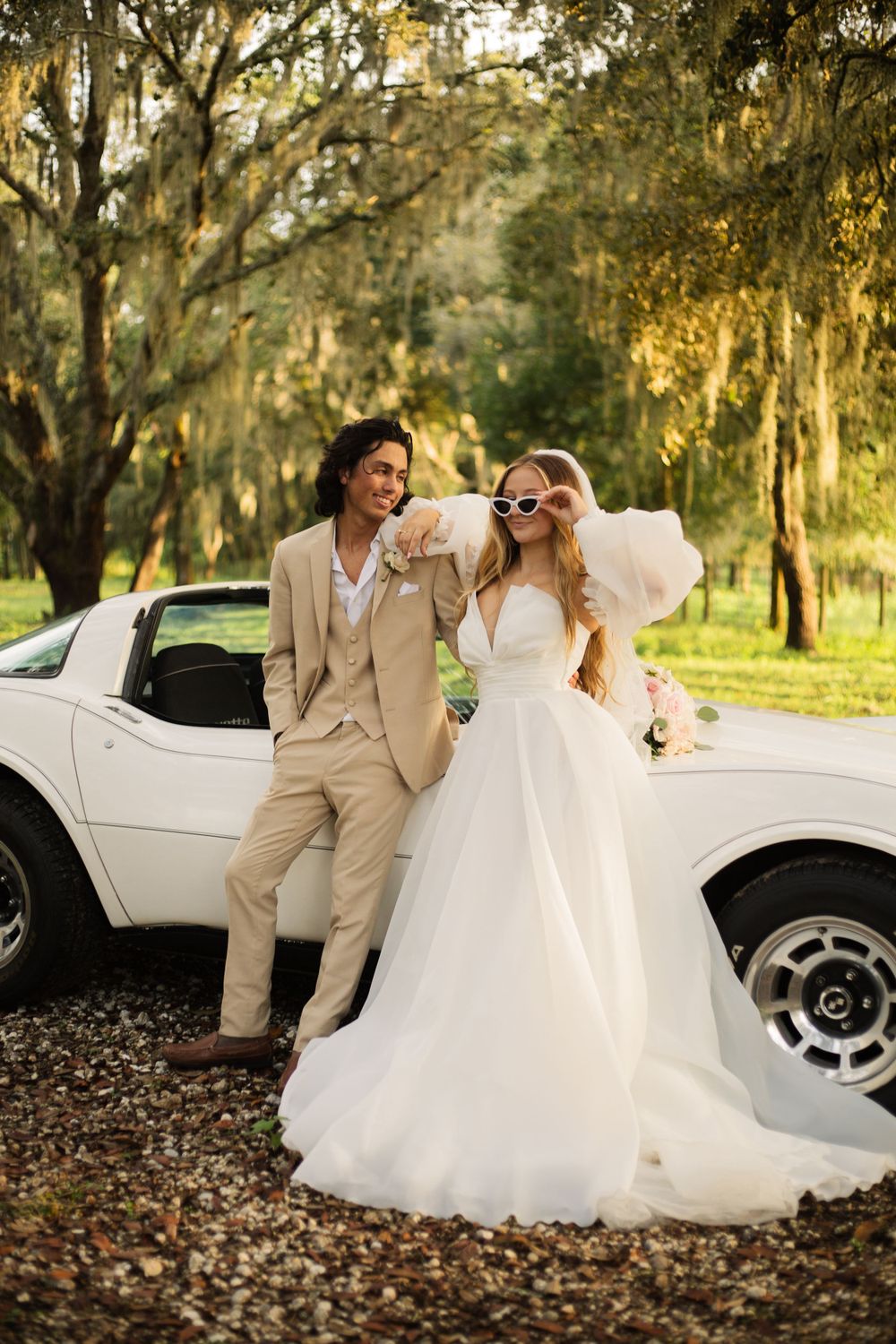 Playful wedding portrait with couple posing against white Corvette under Spanish moss trees.