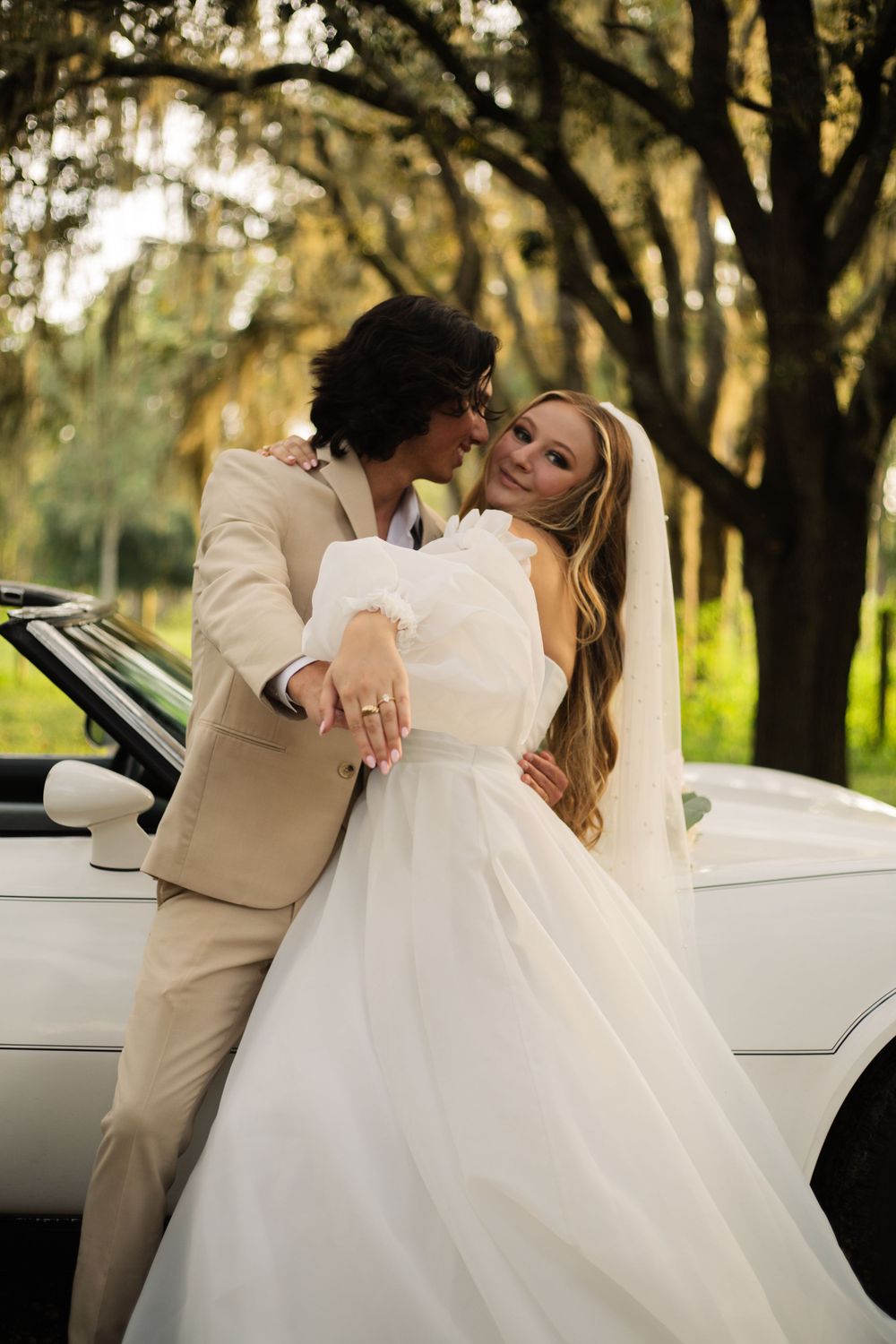 Elegant wedding portrait against vintage white Corvette in a dreamy oak-lined setting.
