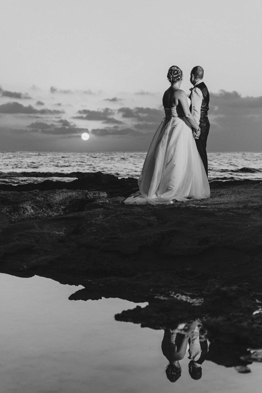 A silhouetted couple stands on rocky coastline at sunset with their reflection in a tidal pool.