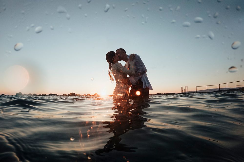 A romantic couple embraces in ocean water during sunset with water droplets in the foreground.