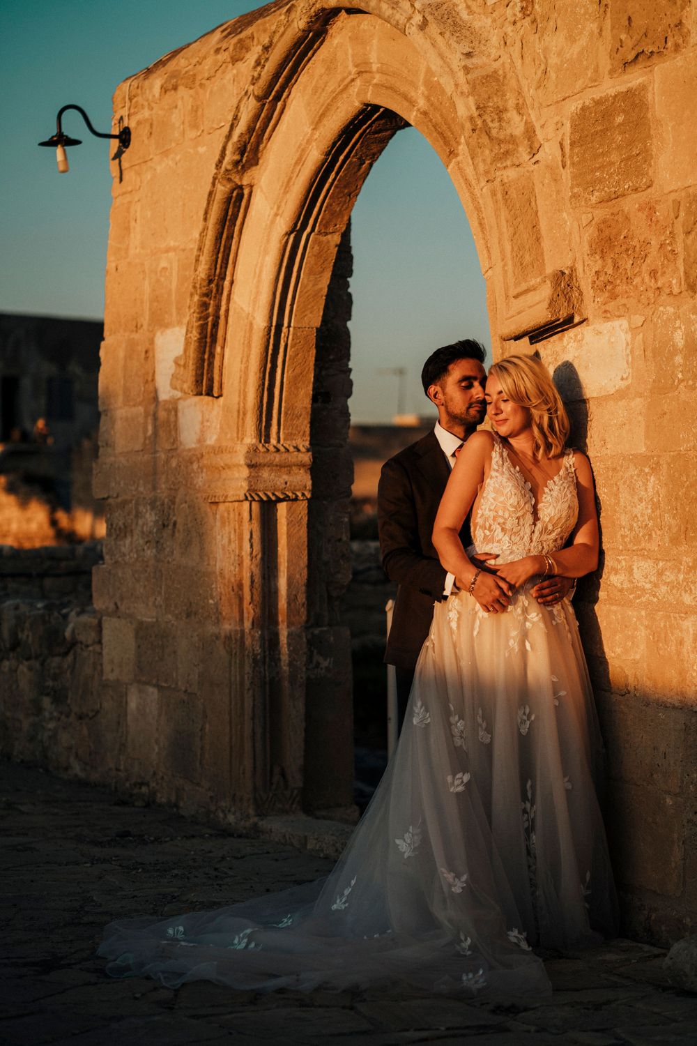 A wedding couple poses beneath a historic stone archway during golden hour.