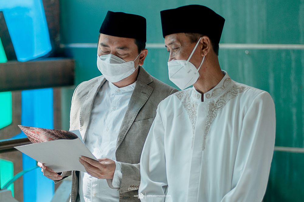 Two men in traditional attire and masks reviewing documents.