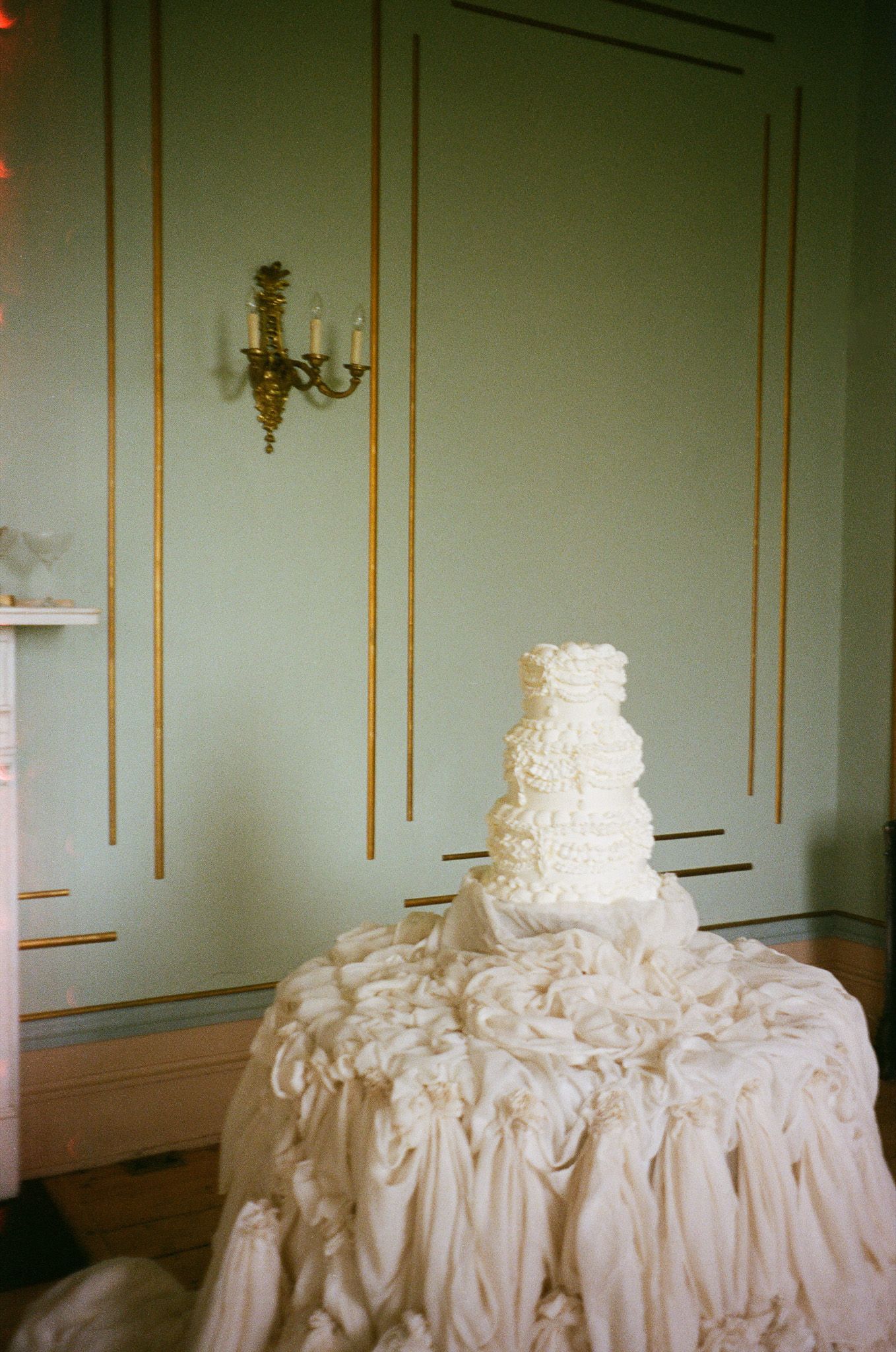 Elegant white wedding cake displayed on elaborately draped table with gold-trimmed mint walls and crystal sconce in background.