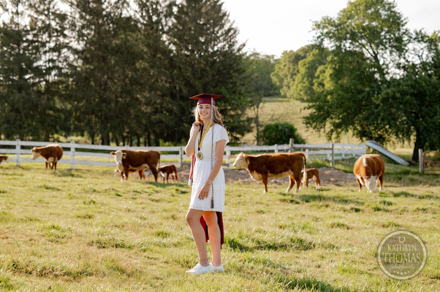 Graduate stands in white dress among grazing cattle in pastoral farm setting.