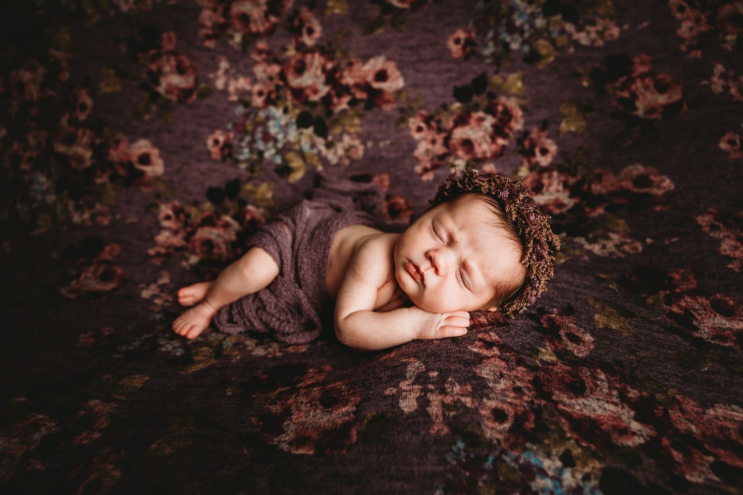 Peaceful sleeping newborn baby wrapped in purple floral blanket with delicate flower headband.