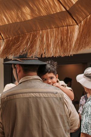 A child peering over someone's shoulder under a thatched roof canopy at an outdoor event.