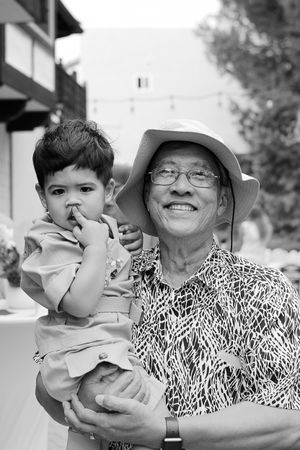 A heartwarming black and white portrait of a young child being held by an elder wearing a sun hat and patterned shirt.