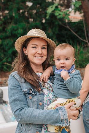 An outdoor gathering showing someone in a denim jacket and straw hat holding a baby in blue clothing.
