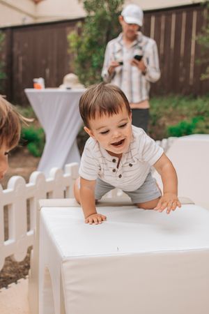 A toddler in a white henley shirt climbing on white furniture at an outdoor party.