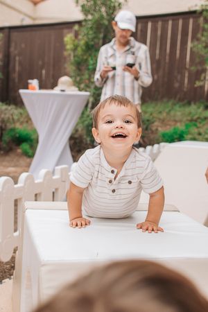 A candid shot of a laughing toddler climbing on white furniture at an outdoor event.