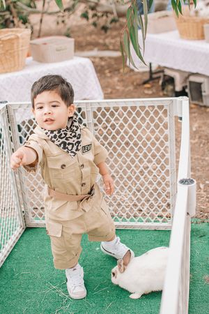 A child in safari-style clothing bends down to pet a white rabbit on artificial grass.