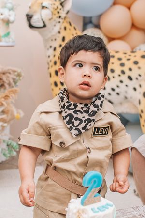 A toddler in a tan safari outfit and leopard print bib celebrates their birthday with cake and balloons.