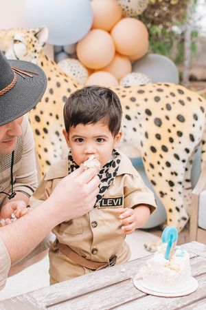 A small child in khaki outfit enjoys birthday cake while surrounded by peach and animal print party decorations.