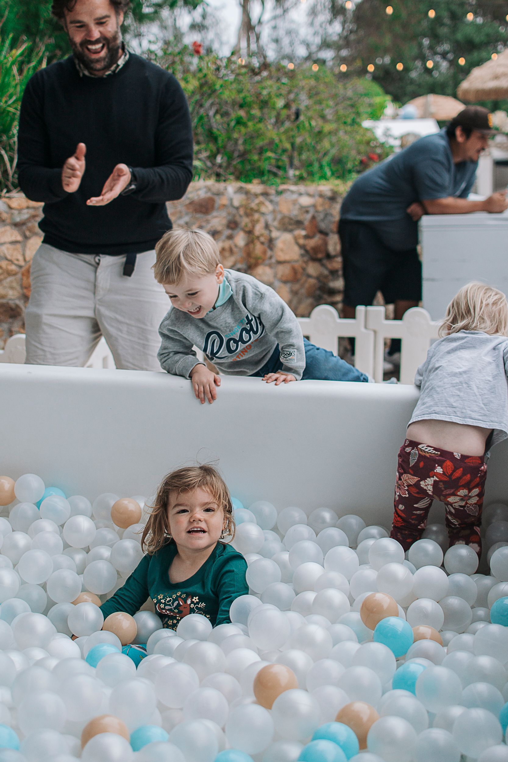 Children playing in a white ball pit filled with blue and beige balls at an outdoor party.