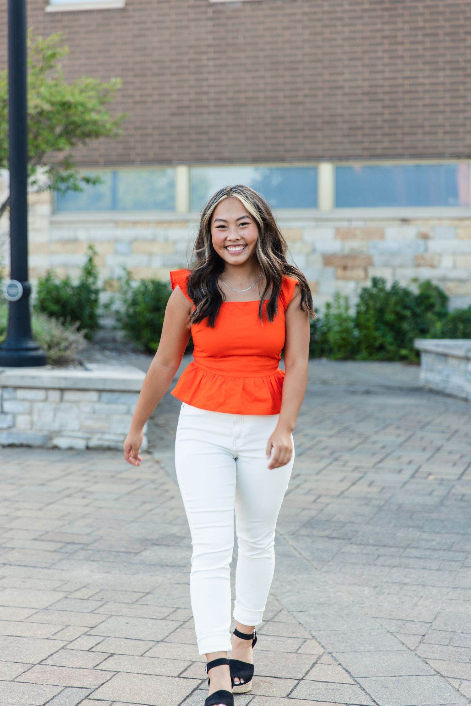 A woman in a bright orange sleeveless top and white pants poses outdoors on a stone paved walkway.