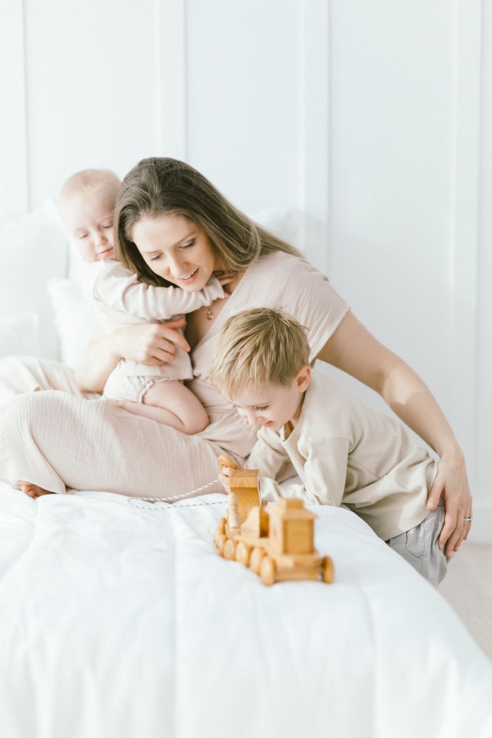 Family playtime with wooden toys on white bedding in bright natural lighting.