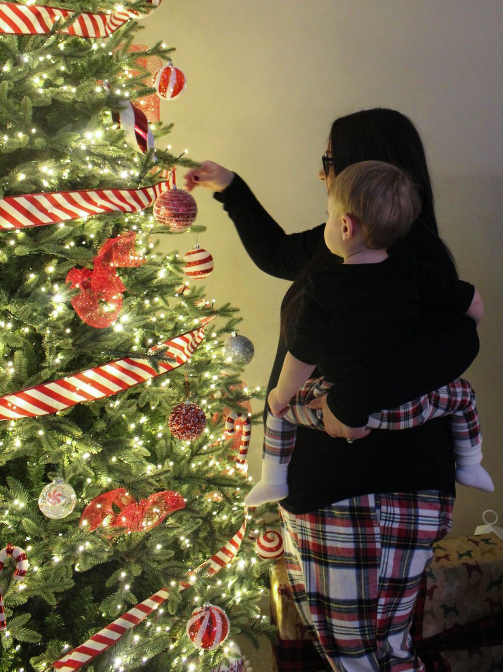 Person in plaid pajamas holds child while decorating Christmas tree with candy cane ornaments.