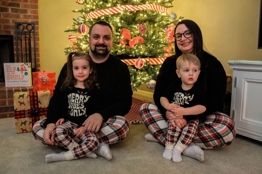 Holiday family portrait with matching plaid pajamas in front of decorated Christmas tree and fireplace.