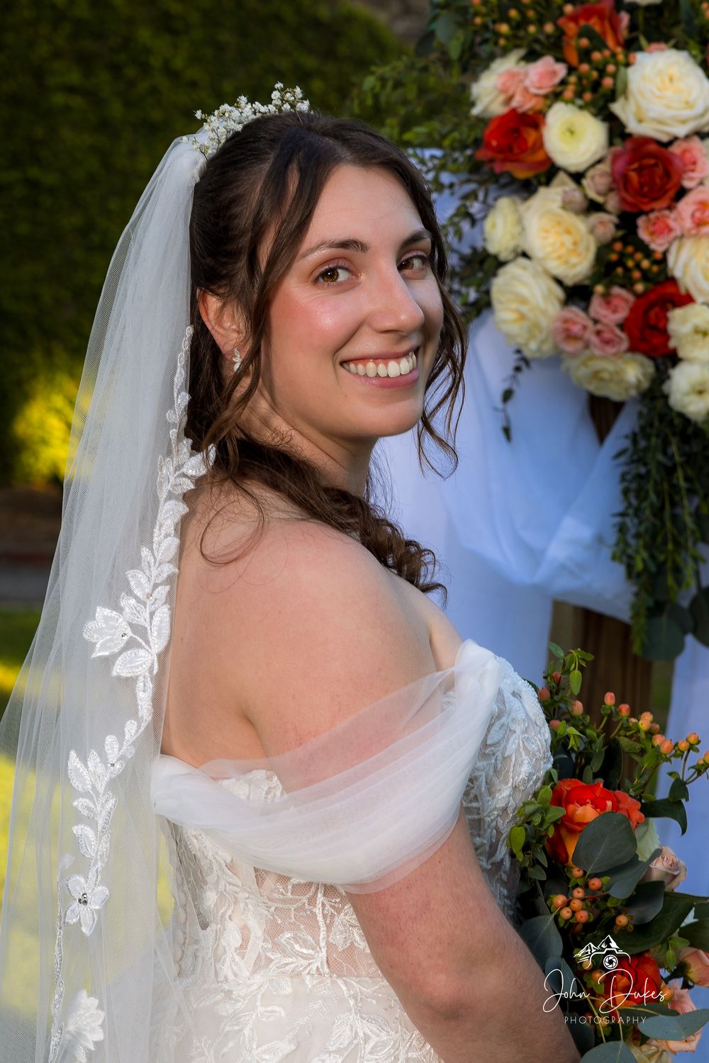 A bride in a white lace dress with veil poses next to colorful floral arrangements at an outdoor wedding.