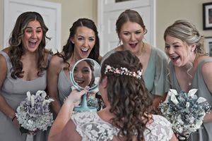 Bridesmaids in gray dresses react with excitement during a wedding day moment.