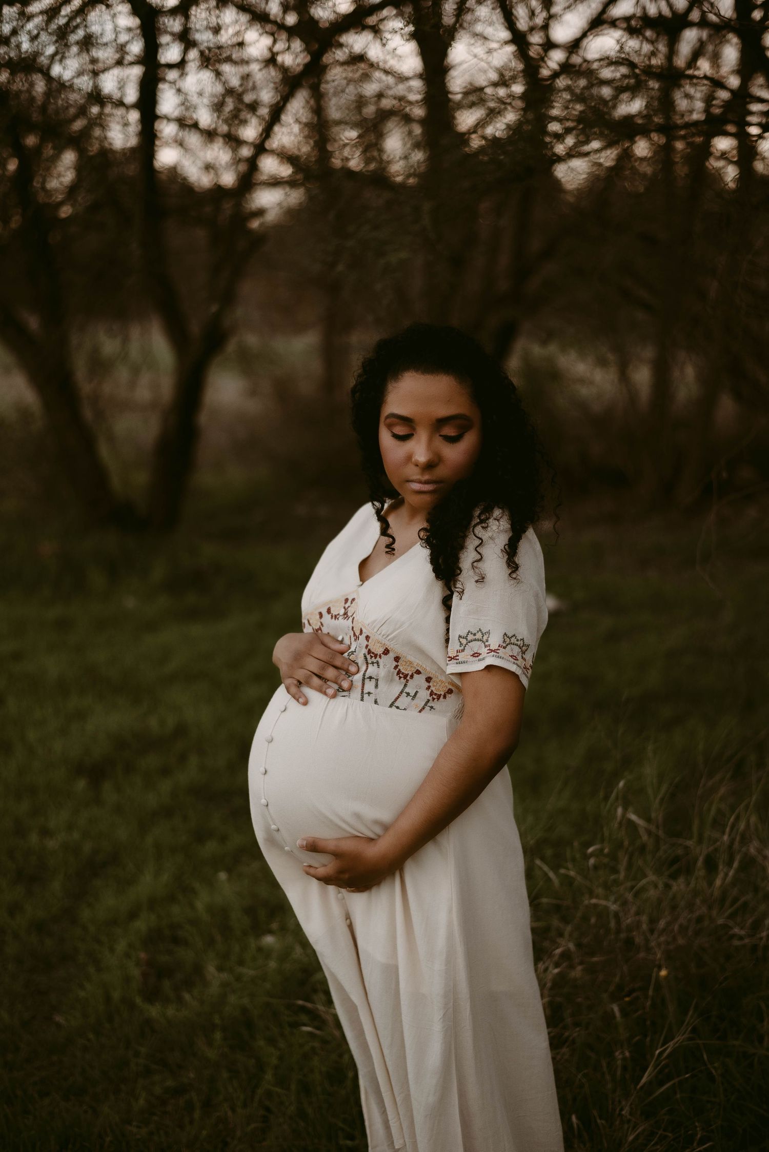 A maternity portrait in a white dress with embroidered details photographed at dusk in a natural setting.
