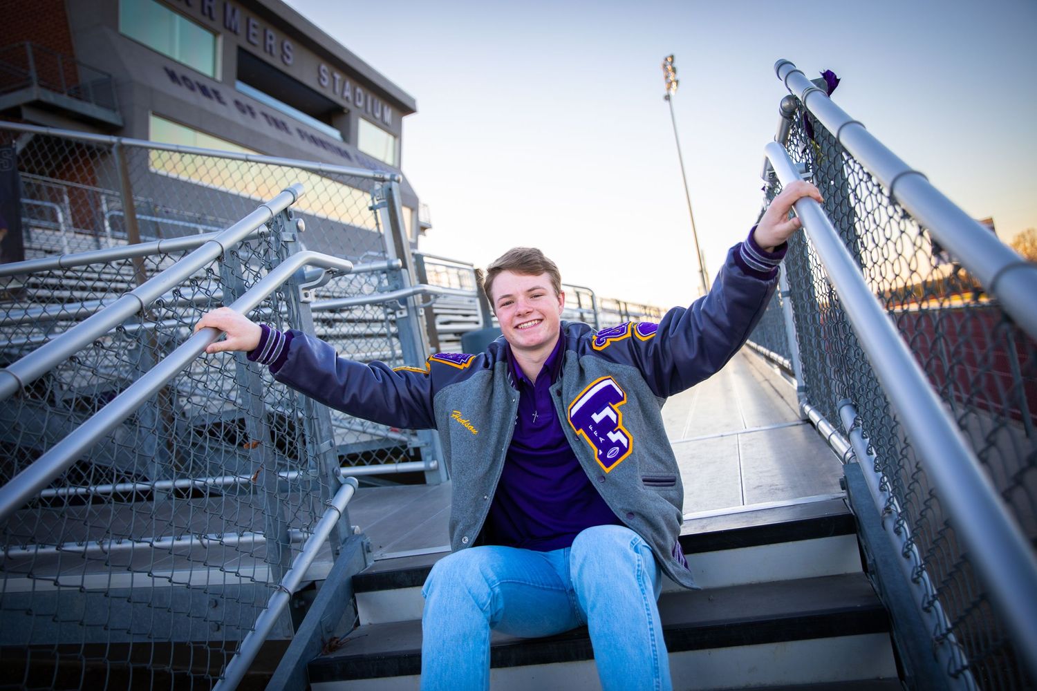 A person in a varsity jacket sits joyfully on metal stadium bleachers with arms raised against a blue sky.