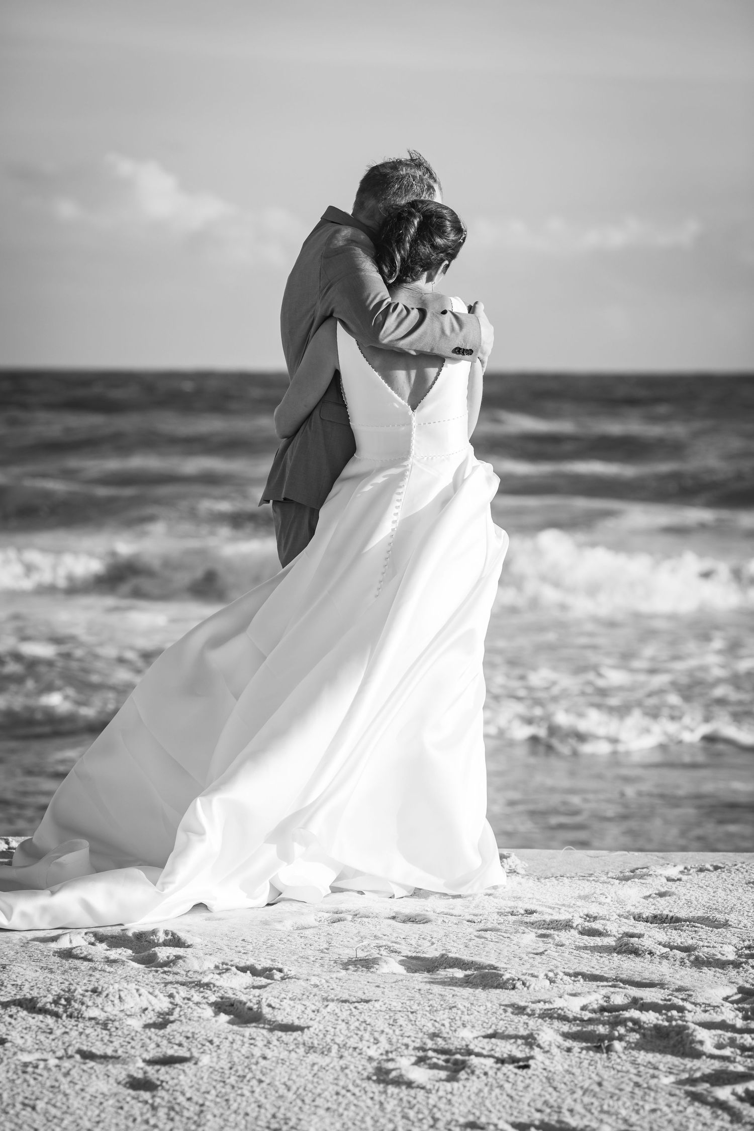 A romantic black and white beach wedding photo of a couple embracing as waves crash behind them.