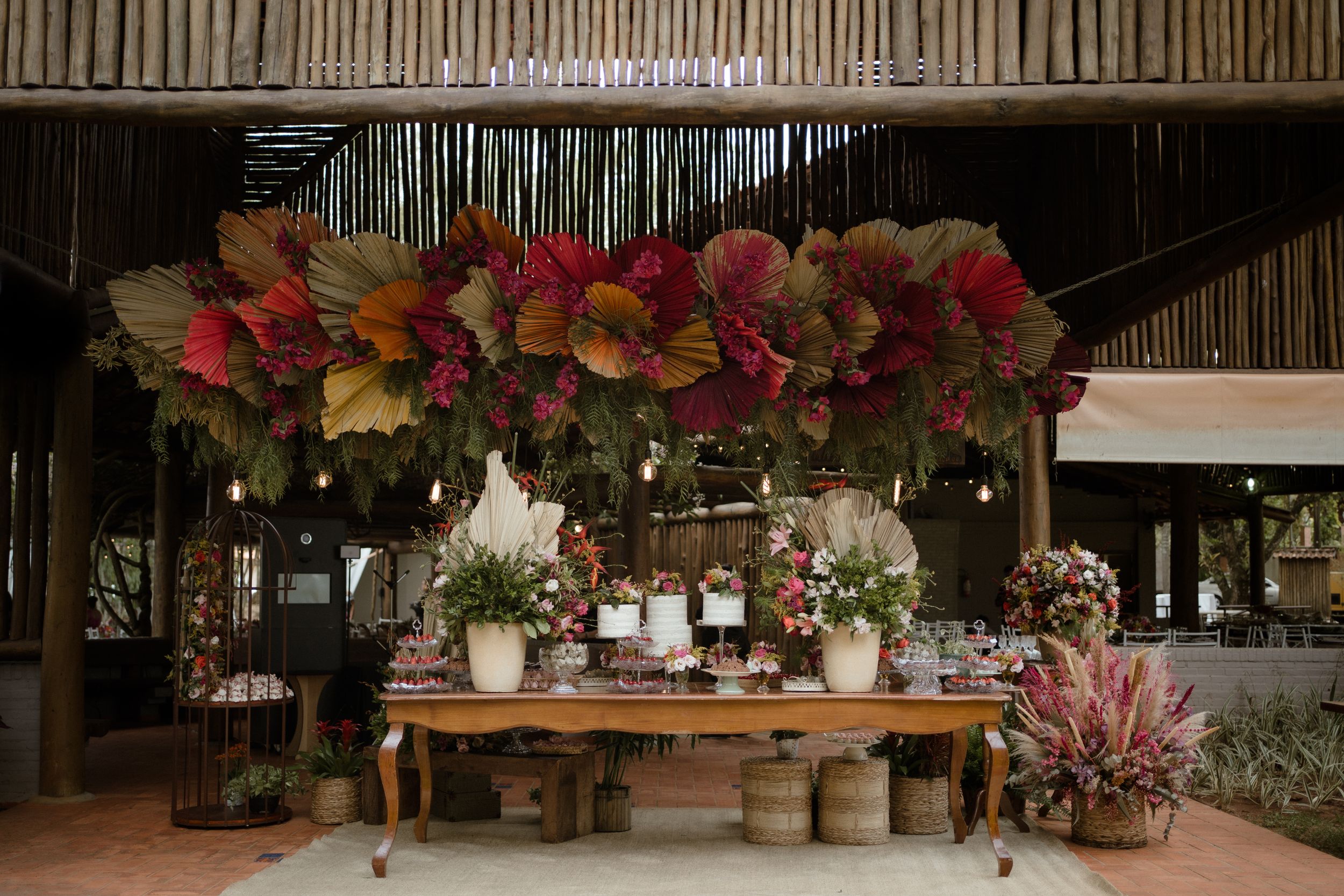 Rustic dessert table with vibrant floral arrangements and bamboo decor overhead.