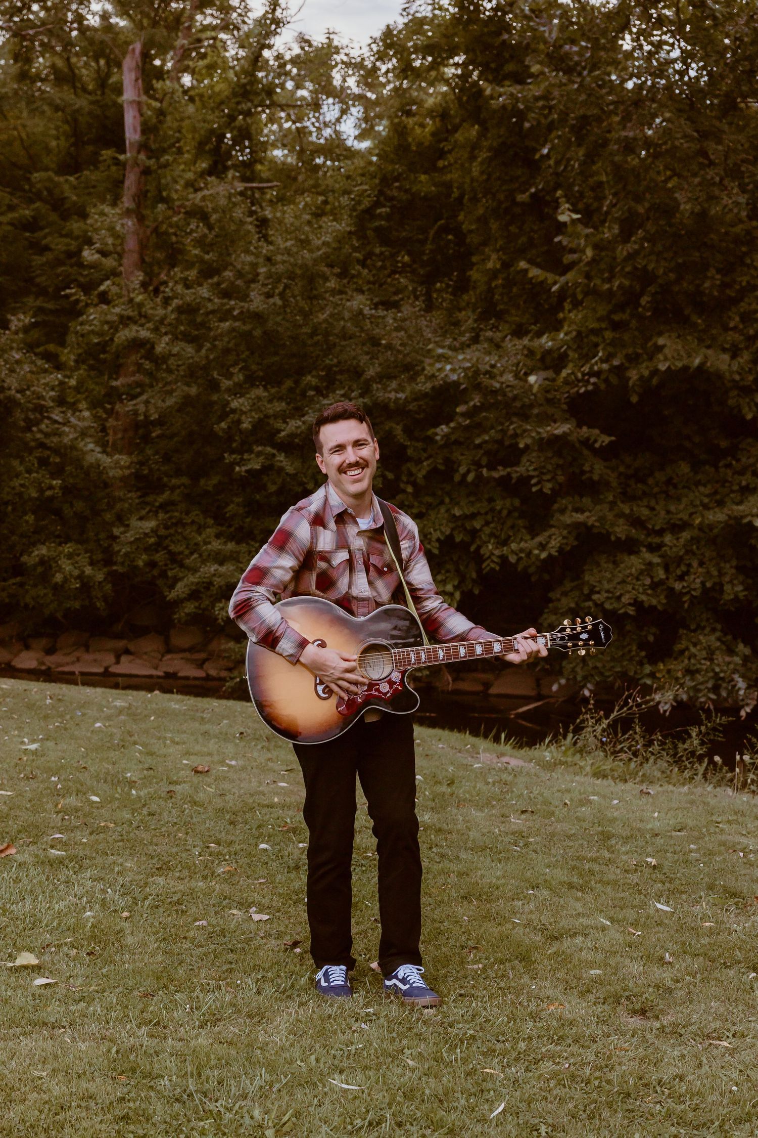 Person playing acoustic guitar while standing in a park setting.
