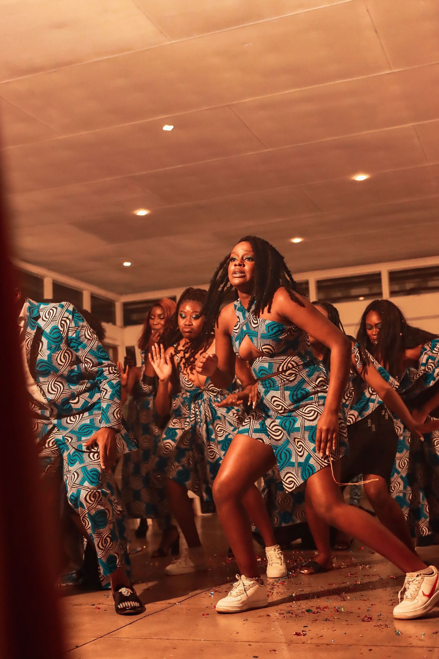 Dancers in vibrant blue patterned dresses perform energetic moves at an indoor celebration.